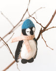 Felt penguin ornament with a blue hat and pink scarf hanging on bare branches.