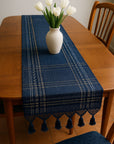 Blue patterned table runner with tassels on a wooden table, featuring a white vase with tulips.