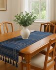 Dining room with a wooden table, blue plaid table runner, white vase with greenery, and wooden chairs.