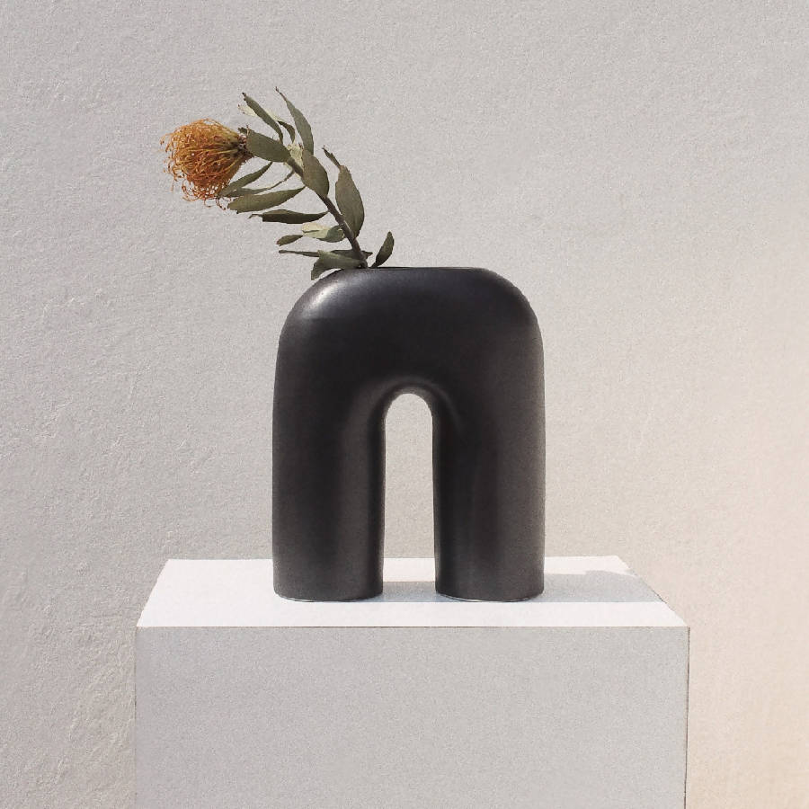 A black, sculptural, inverted-U-shaped vase displayed on a white pedestal with a small floral arrangement on top.