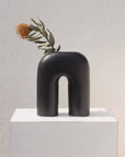 A black, sculptural, inverted-U-shaped vase displayed on a white pedestal with a small floral arrangement on top.