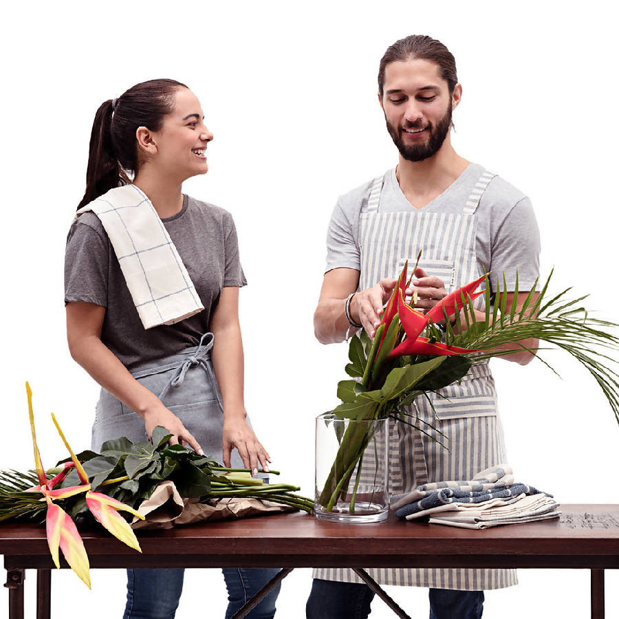 Two people wearing aprons. One is wearing a striped crossback bib apron with multiple pockets and a utility loop. The other is wearing a waist apron.