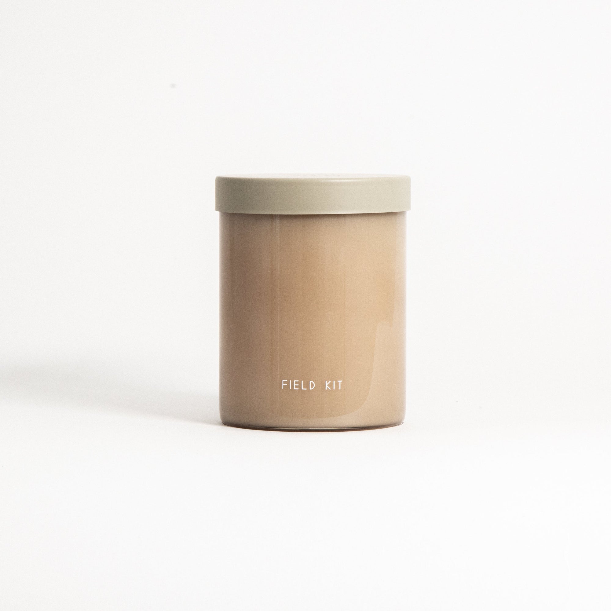 A beige candle in a clear glass jar with a gray lid, labeled 'FIELD KIT', on a light background.