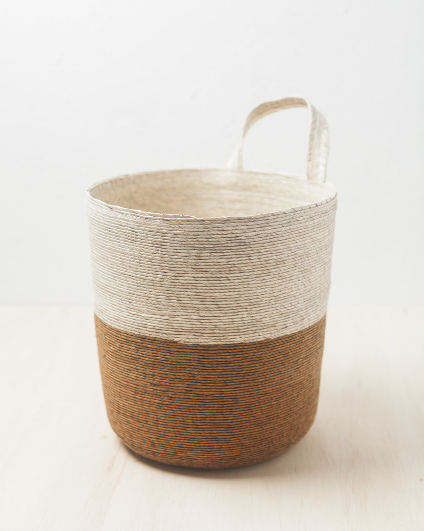A woven basket in a two-tone color, with a natural palm upper section and a brown earthy lower section, displayed against a white background