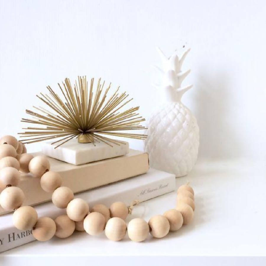 A beige wooden bead garland with jute tassels on each end, displayed alongside a white decorative pineapple and a stack of white books