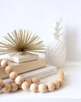 A beige wooden bead garland with jute tassels on each end, displayed alongside a white decorative pineapple and a stack of white books