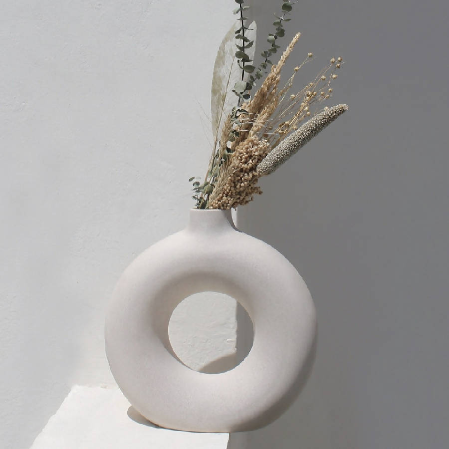 A white ceramic vase with an off-white color and a simplistic, modern design, containing some dried plants, placed against a neutral background.