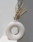 A white ceramic vase with an off-white color and a simplistic, modern design, containing some dried plants, placed against a neutral background.