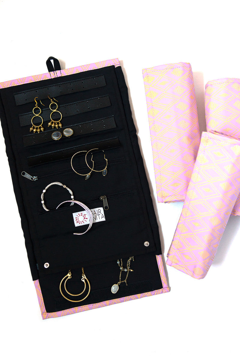 A jewelry roll travel case with various compartments for earrings, rings, and necklaces, displayed alongside some jewelry items and a patterned fabric in pink.