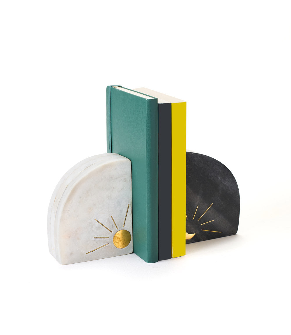 A pair of marble bookends featuring one with a sun design and the other with a moon design, in black, white, and brass colors, with books positioned between them.