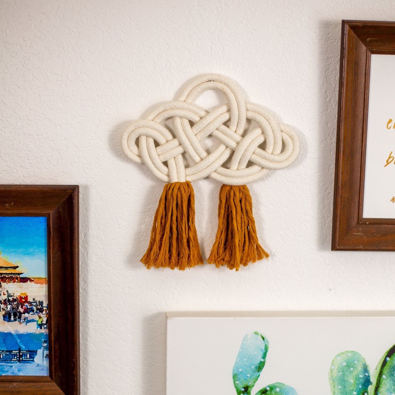 Decorative cotton wall art with braided design and tassels on a wall next to framed pictures.