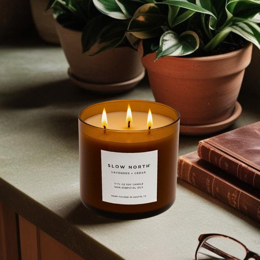 Candle with 'Slow North' label on a surface with potted plants and books in the background