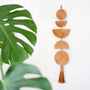 Decorative wall hanging with geometric moon shapes and tassels next to a green leaf on a white background