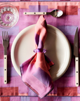 Table setting with a purple and pink checkered placemat, white plate, cutlery, and a folded napkin.