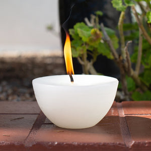 White candle with a single flame on a wooden surface with plants in the background