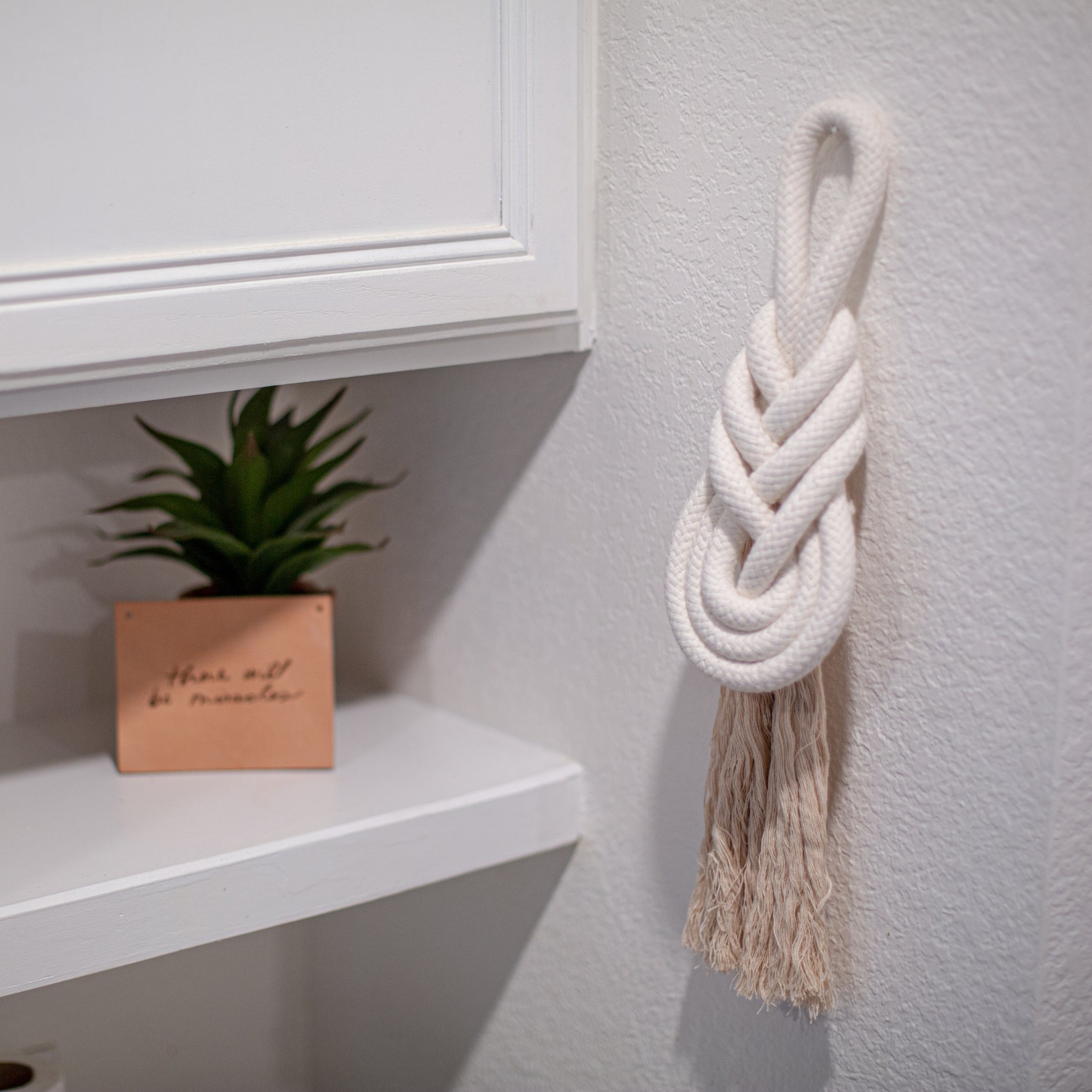 White braided wall hanging on a light gray wall with a plant and shelf in the background