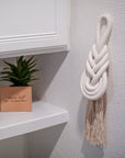 White braided wall hanging on a light gray wall with a plant and shelf in the background
