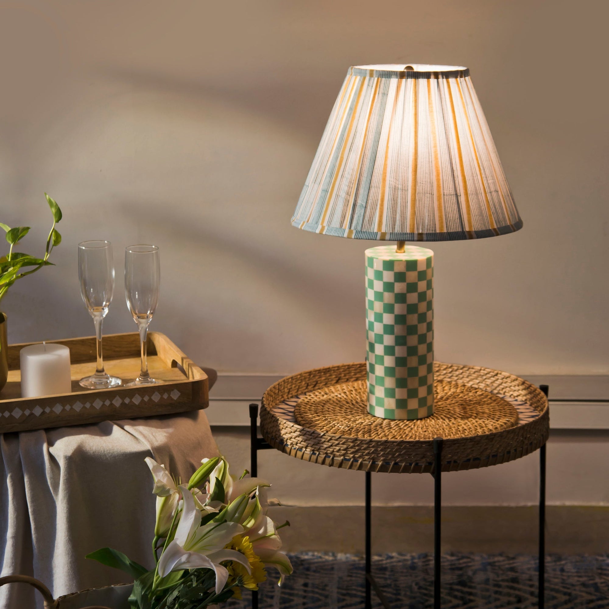 Table lamp with checkered base on a woven side table, with glasses and a plant in the background.