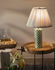 Table lamp with checkered base on a woven side table, with glasses and a plant in the background.