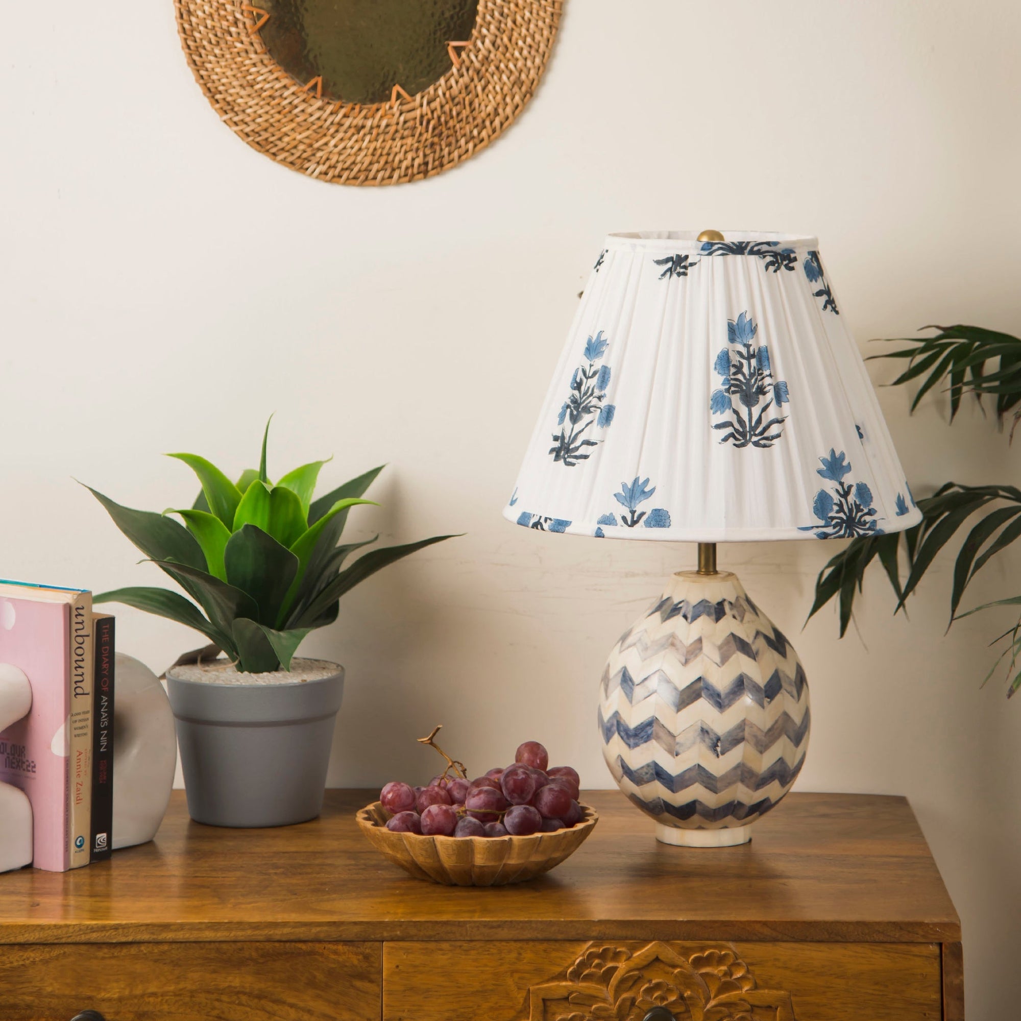 Decorative lamp with floral shade on a wooden table with books, grapes, and a plant.