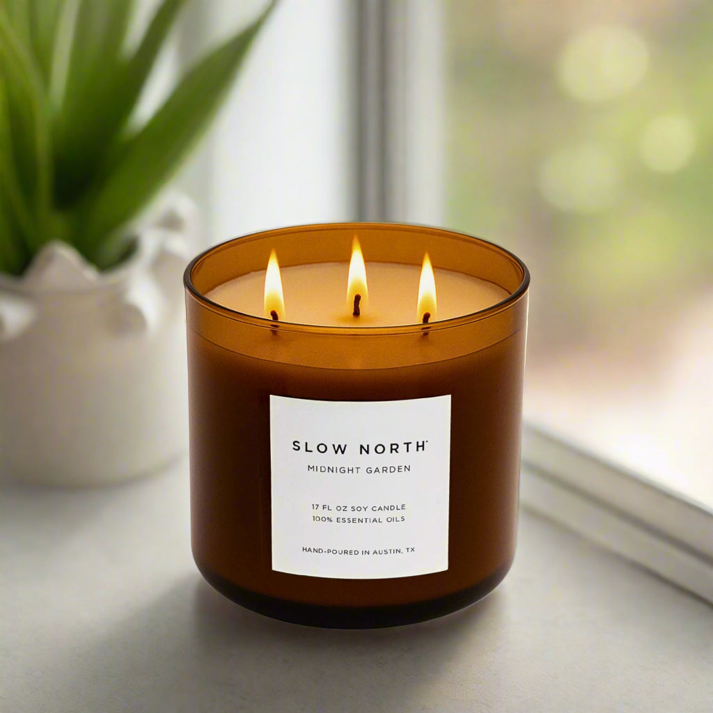 Three-wick candle labeled 'Slow North' on a surface with a blurred natural background