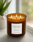 Three-wick candle labeled 'Slow North' on a surface with a blurred natural background