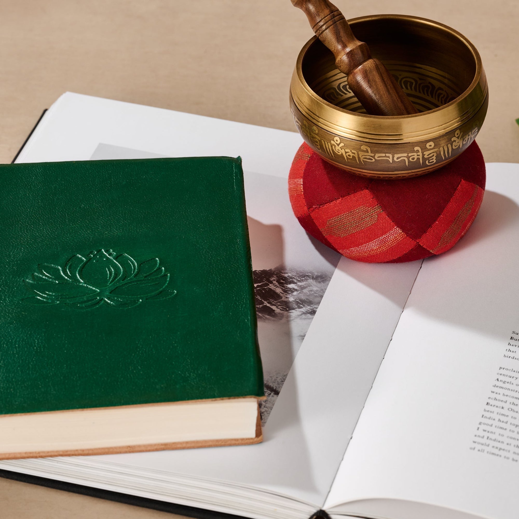 Green book with a lotus embossment next to a bronze singing bowl on a red cushion.