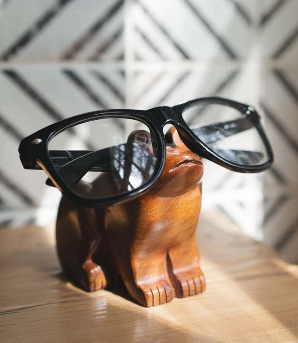 Wooden frog-shaped glasses holder with black-framed glasses on a wooden surface.