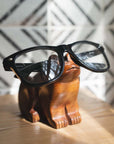 Wooden frog-shaped glasses holder with black-framed glasses on a wooden surface.
