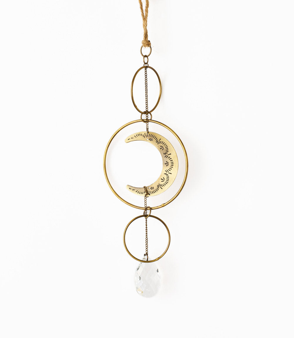 A brass suncatcher with a crescent moon design, consisting of gold-colored metal rings and a white crystal pendant, hanging against a white background.
