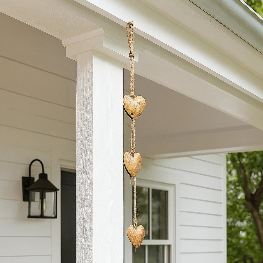 Three hanging wooden hearts in a vertical string decoration, displayed on a white porch with a window, wall sconce, and greenery in the background.