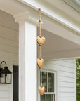 Three hanging wooden hearts in a vertical string decoration, displayed on a white porch with a window, wall sconce, and greenery in the background.