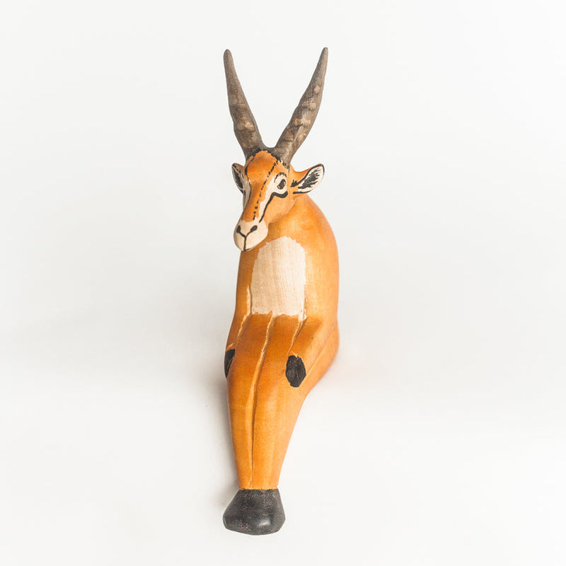 A handcrafted wooden antelope figurine with long curved horns, painted in natural light brown and orange tones with white and black details, set against a plain white background.