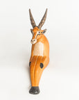 A handcrafted wooden antelope figurine with long curved horns, painted in natural light brown and orange tones with white and black details, set against a plain white background.