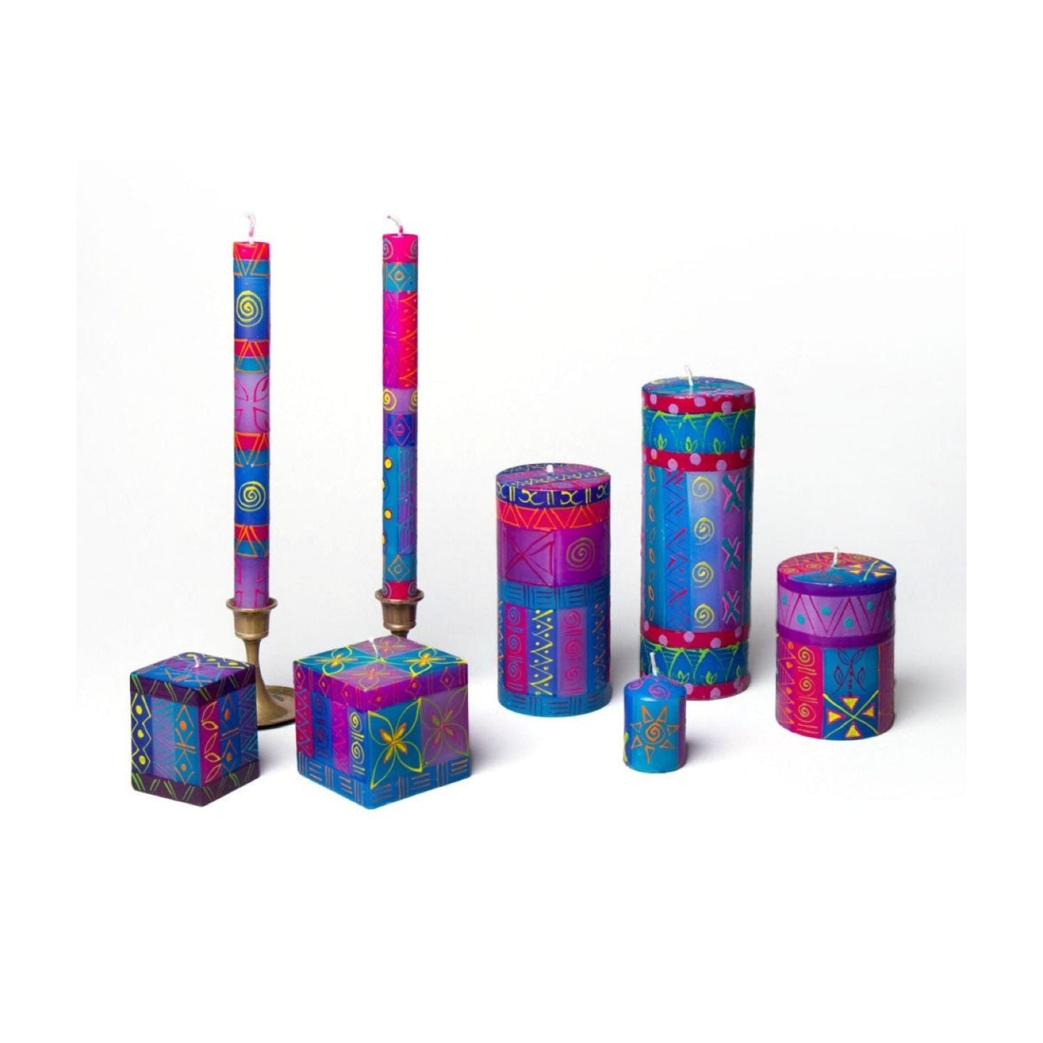 A set of colorful patterned candles in various shapes and sizes, including two tall taper candles on brass holders, a cube candle, a cylindrical candle, a small jar candle, and a round tin candle, all decorated with vibrant geometric and moon motifs in blue, purple, pink, and yellow on a white background.