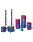 A set of colorful patterned candles in various shapes and sizes, including two tall taper candles on brass holders, a cube candle, a cylindrical candle, a small jar candle, and a round tin candle, all decorated with vibrant geometric and moon motifs in blue, purple, pink, and yellow on a white background.