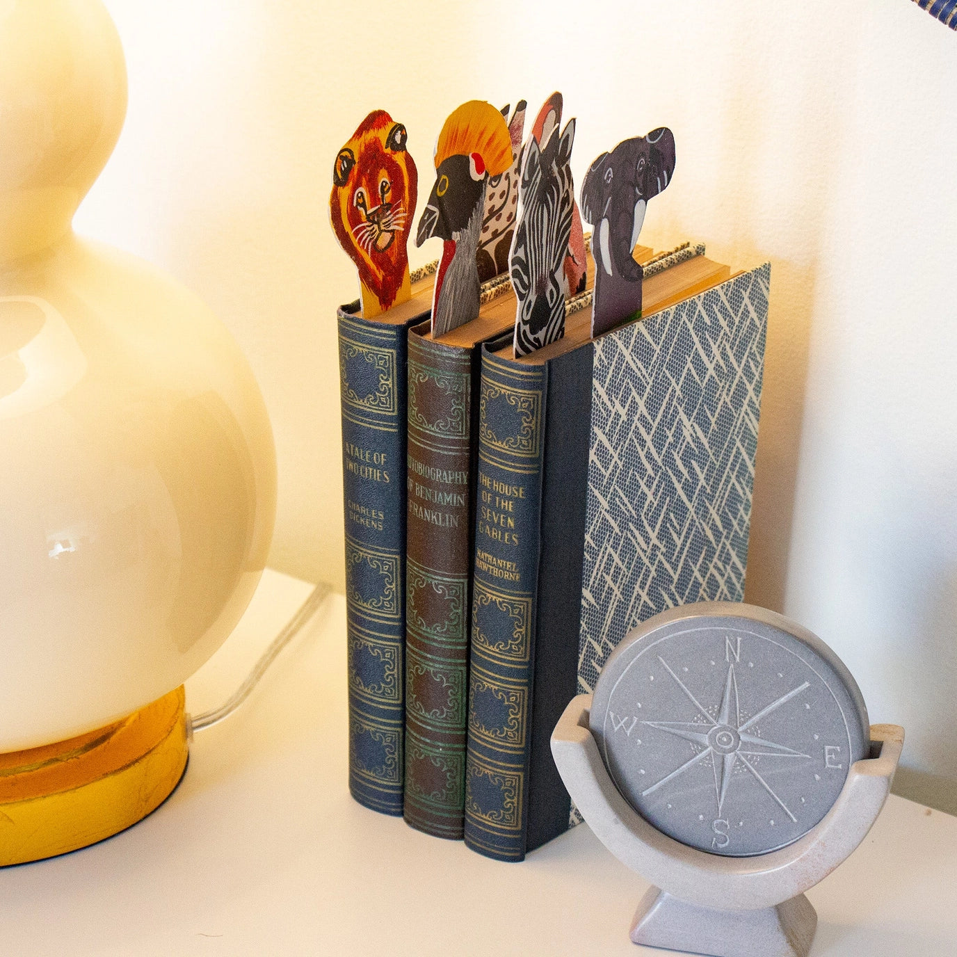 Decorative bookmarks shaped like animals inside of books with a lamp and compass in the background.