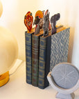 Decorative bookmarks shaped like animals inside of books with a lamp and compass in the background.