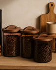 Wooden canisters with coffee beans on a wooden surface with a coffee maker and mug in the background.
