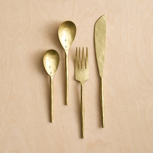 A set of four gold-colored flatware pieces laid out on a wooden surface, including a fork, knife, soup spoon, and teaspoon