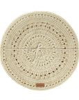 A beige, round, handwoven placemat with a spiral pattern and a label indicating the brand at the bottom