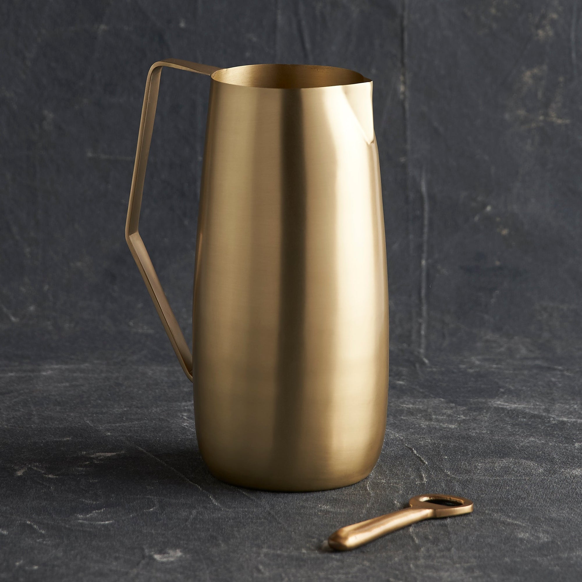 A hand-crafted brass pitcher with a shiny finish, featuring an angled handle and a pinched spout, displayed next to a key-shaped tool.