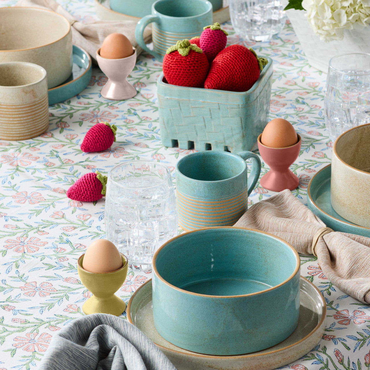 Table setting with ceramic bowls, cups, and decorative eggs on a floral tablecloth.