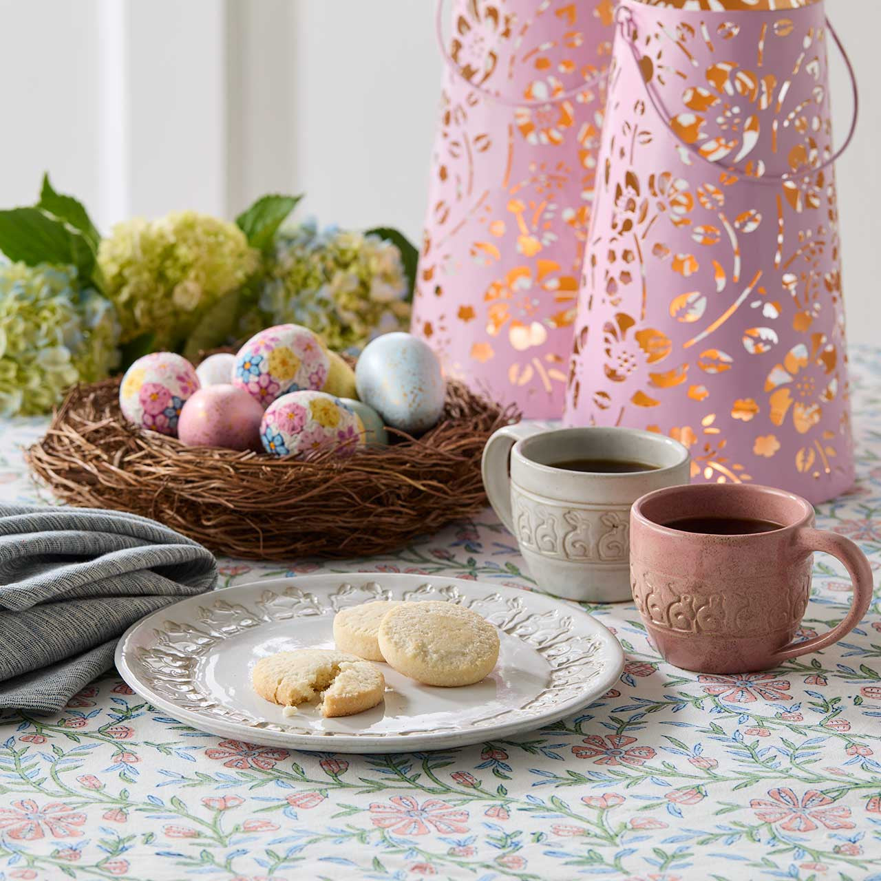 Easter eggs in a nest with floral patterns, two mugs, and cookies on a tablecloth.