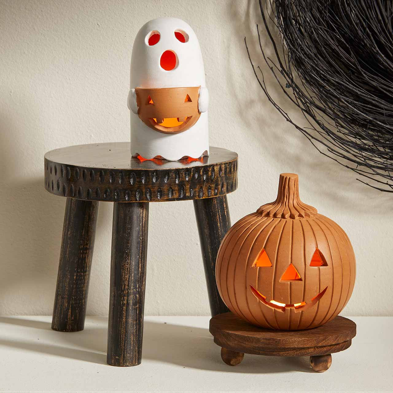A Halloween-themed tabletop display featuring a white ghost-shaped candle holder with a glowing orange pumpkin face, set on a small dark wooden stool, and an orange jack-o'-lantern candle holder on a round wooden base in front of it.