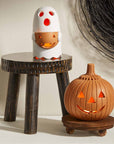 A Halloween-themed tabletop display featuring a white ghost-shaped candle holder with a glowing orange pumpkin face, set on a small dark wooden stool, and an orange jack-o'-lantern candle holder on a round wooden base in front of it.