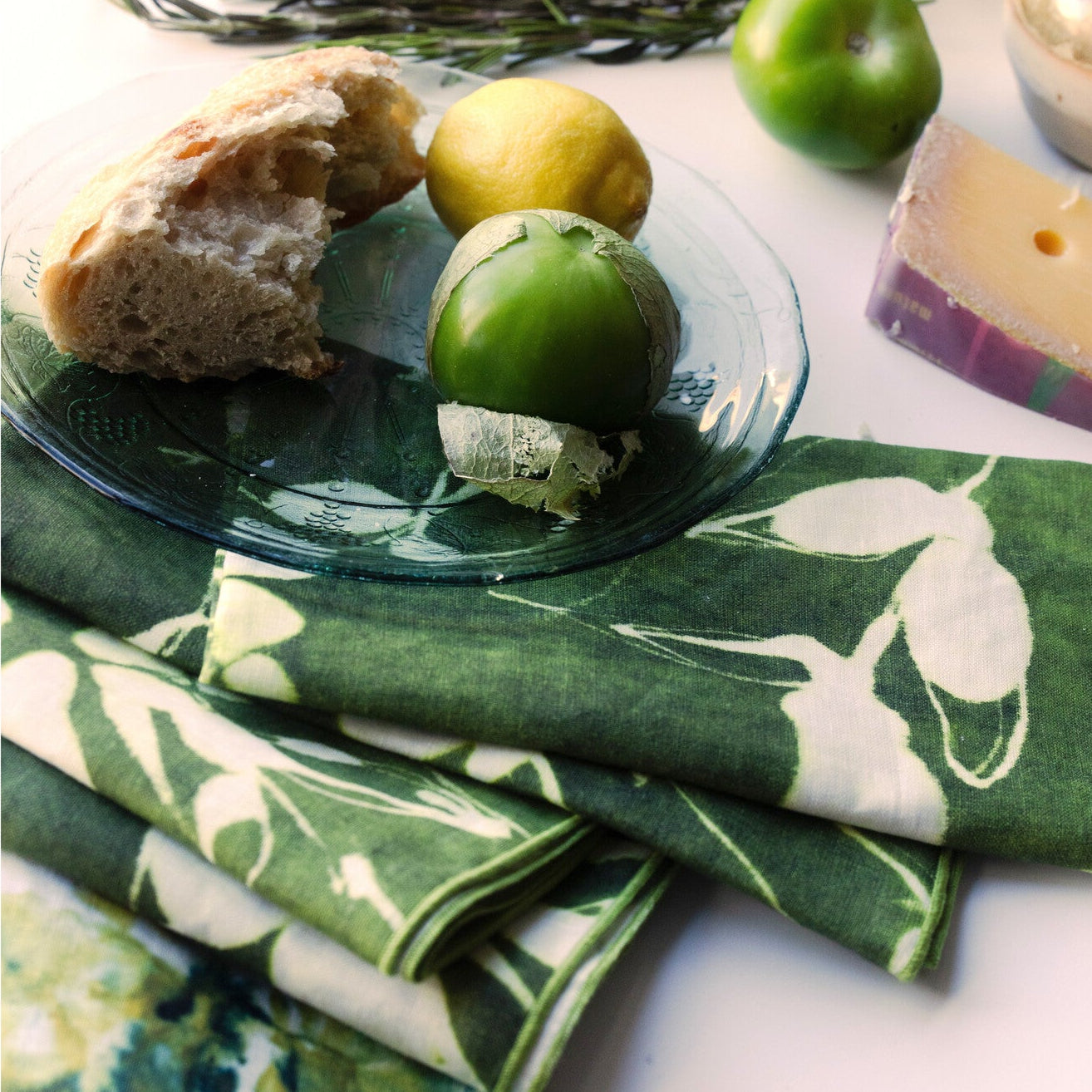 Green floral-patterned fabric with a glass dish containing bread, fruit, and cheese on a white surface.