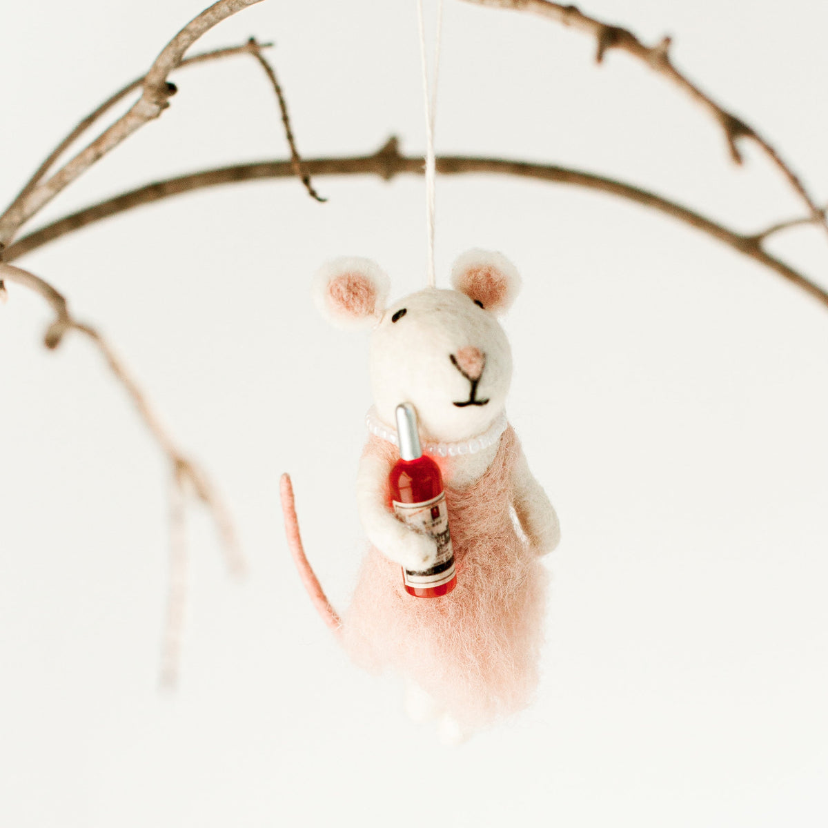 A felted wool mouse ornament wearing a pink dress and holding a small red bottle, hanging from a thin branch against a clean white background.