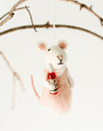 A felted wool mouse ornament wearing a pink dress and holding a small red bottle, hanging from a thin branch against a clean white background.