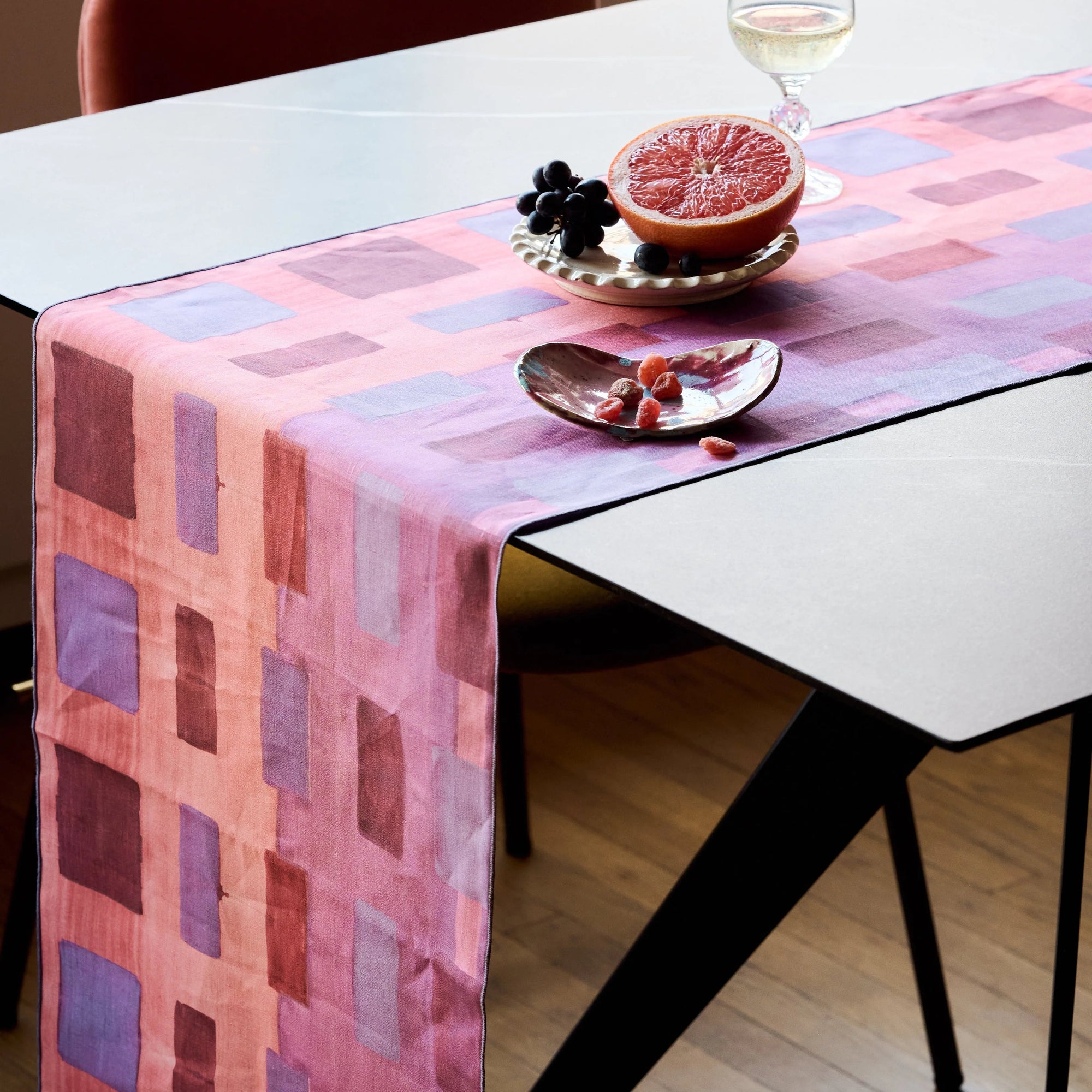 Table with a pink and purple checkered tablecloth, fruit on a plate, and a glass of white wine.